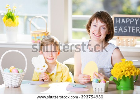 happy smiling family of two enjoying easter time at home, doing crafts, little boy holding easter bunny shaped candy, easter themed decorations and blackboard sign "happy easter!" in the background