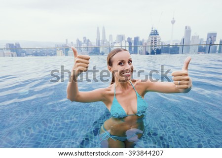Vacation in Kuala-Lumpur. Pretty young woman showing thumbs up while swimming in roof top pool with beautiful city view.