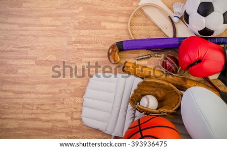 sports equipment on a gym floor, football, hockey stick, rugby ball, baseball, cricket bat and pads, basketball, boxing gloves, badminton and squash racket. Royalty-Free Stock Photo #393936475