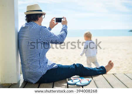 Rear side view of father with smartphone taking picture of baby. beach terrace on sunny day outside background