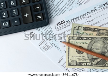 1040 Individual Income Tax Return Form for 2015 year with a pencil to fill in, calculator and dollar bills on the white desk, top view