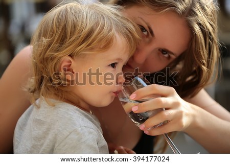 Smiling young mother holding glass and child boy with blonde hair drinking water from paret hands sunny day outdoor, horizontal picture