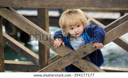 One cute funny little baby boy with blonde hairstyle in blue jacket hiding behind wooden fence outdoor looking forward, horizontal pictures