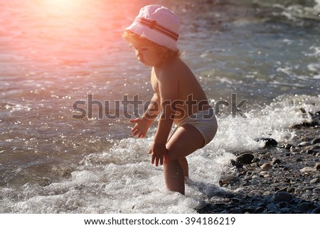 Little curious funny blonde baby boy in hat sitting on sand sunny day outdoor playing with wavy ocean water on natural beach background, horizontal picture