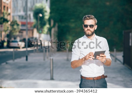 Knee figure of young handsome bearded businessman holding a tablet, looking over wearing sunglasses - technology, business, work concept, copy space left Royalty-Free Stock Photo #394365412