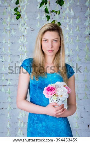 Portrait of beautiful smiling woman in blue dress  standing near garland made of white artificial bell-flowers holding peonies bouquet in a small white vase