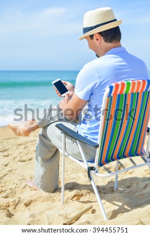 Back side view of young man relaxing on the beach outdoors background using smartphone. Barefoot luxury relaxation
