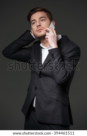 Pensive attractive young businessman in black suit talking on smartphone and thinking  over grey background