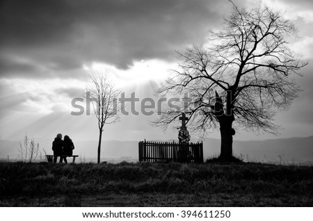 Two young people in love are sitting together on the bench near the tree and cross. They are looking to wonderful spring  sky full of rays.