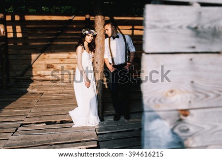 young beautiful couple posing on the background of a wooden house in the direct sunlight. she is in a white dress and a wreath of flowers in her hair, and he with suspenders and a butterfly