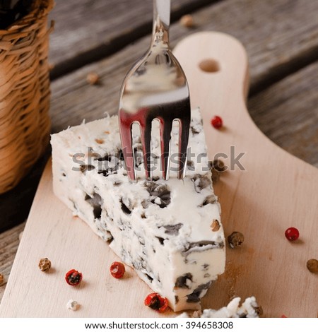 Square photo of  blue cheese portion placed on wooden board with colour pepper seeds around. Bottle of wine is next to cheese and small fork is stuck inside. All is on old grey table.