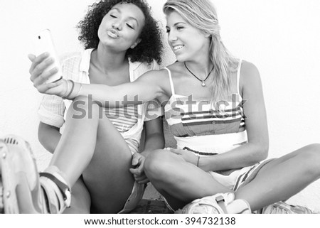 Black and white portrait of ethnically diverse teenager girls friends posing together outdoors, wearing skates using smart phone to take selfies pulling faces. Teenagers active lifestyle technology.