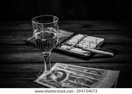 Glass of vodka with money of the Soviet Union and cigarette with cigarette case on an old wooden table. Angle view, image vignetting and the black and white tones