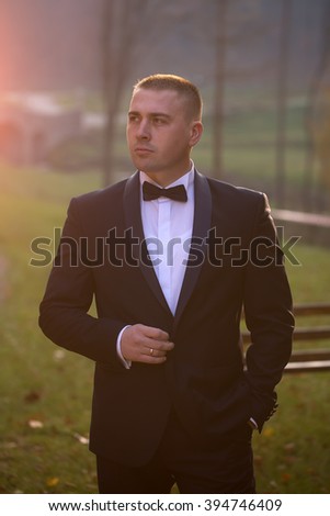 Handsome stylish young bridegroom in black wedding suit jacket white shirt on bow tie standing outdoor in forest on natural background, vertical picture