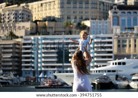 Young mother in  white holding in hads happy smiling blonde chuild boy in port with water transportation and buildings outdoor sunny day, horizontal picture