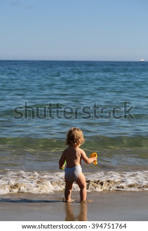 Small curious blonde child boy standing on sea coast beach with wavy water sunny twilight outdoor playing on natural background, vertical picture