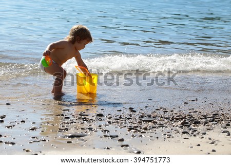 Small curious funny blonde child boy standing on sea coast beach with wavy water sunny day outdoor playing with yellow plastic pail on natural background, horizontal picture