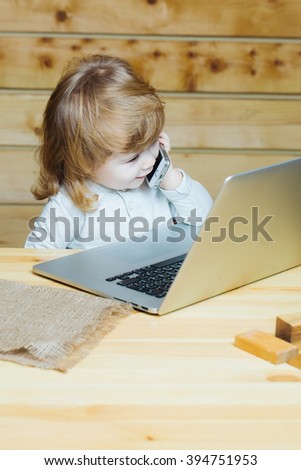 Cute funny little baby boy with long blonde curly hair speaking by mobile phone near computer indoor on wooden background, vertical picture