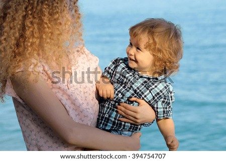 Young pretty woman with long lush curly hair holding happy smiling cute baby boy in hands in checkered shirt outdoor sunny day near sea water on natural background, horizontal picture