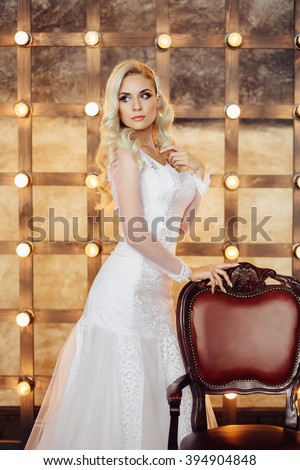 Healthy wedding hair. Attractive bride with long blonde curly hairstyle and bridal makeup standing near the brown chair on background of light bulbs. Beauty indoor close up portrait in white dress.