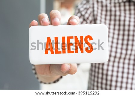 Man hand showing ALIENS word phone with  blur business man wearing plaid shirt.
