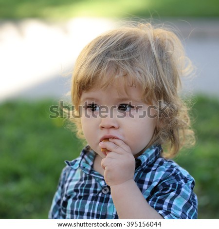 Portrait of cute adorable small serious boy child with blonde hair in stylish blue checkered shirt eating outdoor on summer green grass background, square picture