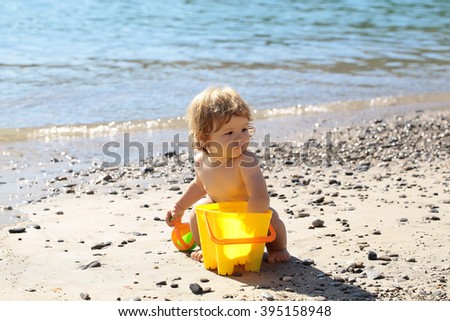 Small curious funny blonde child boy sitting on sea coast beach with wavy water sunny day outdoor playing with yellow plastic pail on natural background, horizontal picture