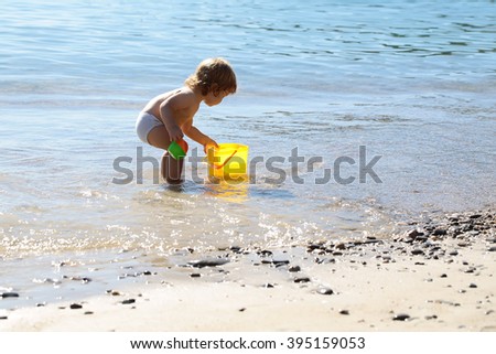Small curious funny blonde child boy standing on sea coast beach with wavy water sunny day outdoor playing with yellow plastic pail on natural background, horizontal picture