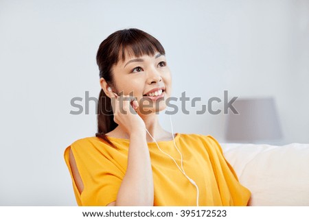 people, technology and leisure concept - happy asian young woman sitting on sofa with earphones listening to music at home