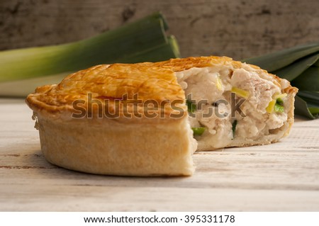 Chicken and leek pie with slice missing on a white wooden surface with fresh leeks in the background. Royalty-Free Stock Photo #395331178