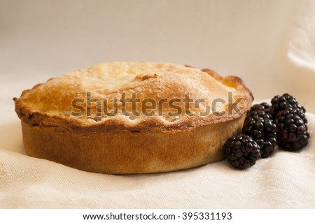 A whole cooked shortcrust pie alongside blackberries on white cloth.
