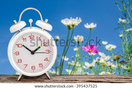 White alarm clock on wooden table with cosmos flower background soft focus.