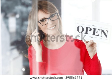 Portrait of middle age small shop owner woman holding in her hand open sign while standing at doorway. 