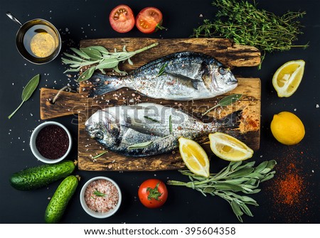 Fresh uncooked dorado or sea bream fish with lemon, herbs, oil, vegetables and spices on rustic wooden board over black backdrop, top view Royalty-Free Stock Photo #395604358