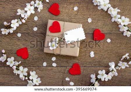 At center gift decorated with red heart, flowering sprig with gift card on, around almond flowering sprigs at corners, 4 hearts, petals on wood background. Spring spirits gift. Horizontal. Top view. 