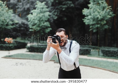 Young handsome beard man in white shirt and trousers makes beautiful pictures on film retro camera, stylishly dressed, photographer,outdoor portrait, close up,brutal, tattoo, street photo,photographer Royalty-Free Stock Photo #395830477