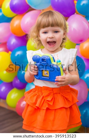 colorful portrait of cute adorable baby girl with balloons background and toy photo camera in her hands