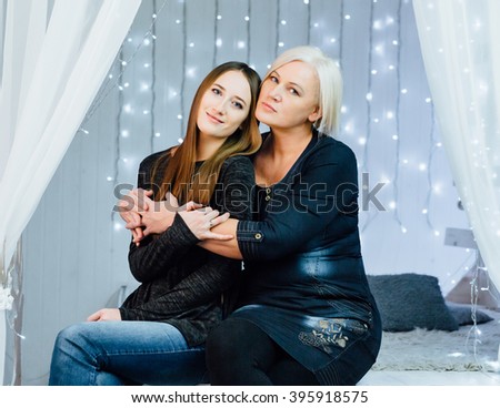 Studio portrait of mother and adult daughter. Mom hugging adult daughter sitting on the couch and looking at the camera.