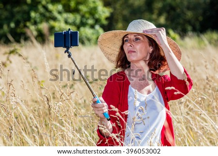 outdoors selfie - smart middle aged woman making a self-portrait on mobile phone on stick in the middle of high dry grass,summer daylight