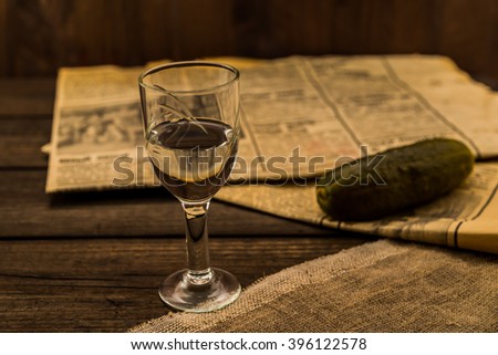 Glass of vodka with newspaper and pickled cucumber with canvas on an old wooden table. Angle view, shallow depth of field, focus on the glass of vodka