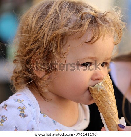 Portrait of small hungry funny baby boy with wet blonde curly hair eating cold tasty ice cream in wafer from hands of mother outdoor, square picture