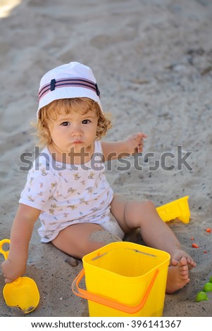 Little curious smart blonde baby boy in hat sitting on sand outdoor playing with plastic colorful toys on natural beach background, vertical picture