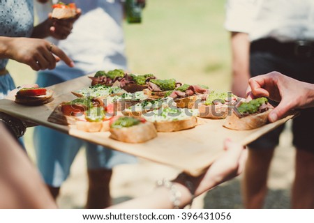 Canapes with avocado, goat cheese and  tomato and people on background. Close-up of hands taking canapes from the tray. 
Snacks for guests on a party. Waiter with the tray and guests around on a event Royalty-Free Stock Photo #396431056