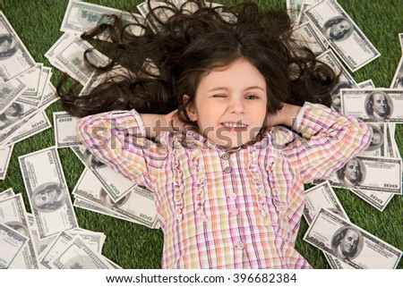 Top view photo of beautiful little girl lying on green grass and dollars. Girl looking at camera, smiling and winking