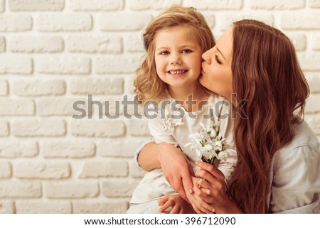 Beautiful young mother is holding flowers and kissing her cute little daughter, against white brick wall. Little girl is smiling
