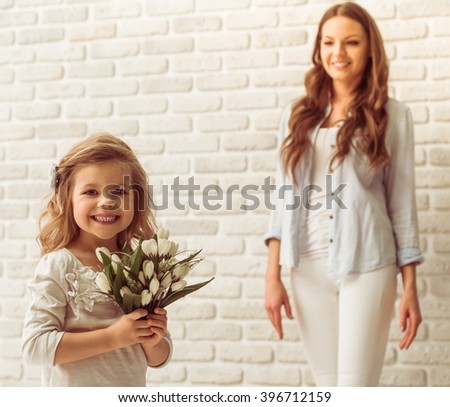 Sweet little girl is holding flowers, looking at camera and smiling, her beautiful young mother in the background
