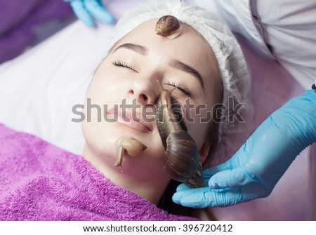 Young woman receiving snail facial massage. Snail on face. Cleaning procedure in spa salon. Blurred background.