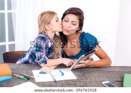Photo of mother and little daughter using tablet computer. Nice white interior with wooden table. Daughter kissing her mother