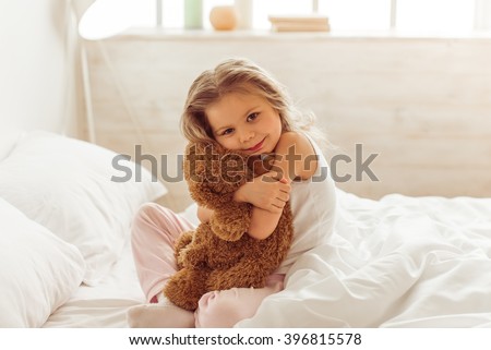 Sweet little girl is hugging a teddy bear, looking at camera and smiling while sitting on her bed at home Royalty-Free Stock Photo #396815578