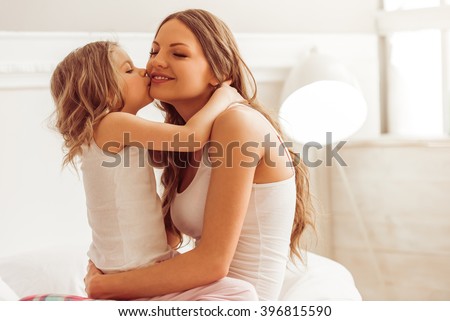Sweet little girl is hugging and kissing her beautiful young mom in cheek while sitting on bed at home